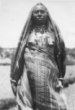 Postava ženy zahalené v rouchu z kostkované látky, s obnaženými prsy a šňůrami korálů na krku, kmen Hamayd
