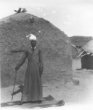 Muž stojící před hliněnou chýší, oblečený v plášti z proužkované látky, na hlavě turban, opírá se o hůl