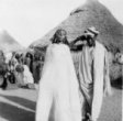 Dívka se šperky ve vlasech, zahalená v bílém přehozu, vedle ní stojí muž v  turbanu, kmen Hamayd, společenství Baggara