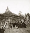 Obytná chýše se slaměnou střechou, u vchodu klečí muž a dvě ženy, Kumam  nebo Teso