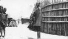 Žena zahalená do přehozu z plátna stojí na dvorku před rákosovou ohradou, Šilukové