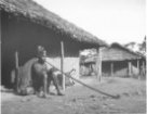Muž sedící na kládě před chatou kouří dlouhou dýmku, Babira