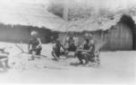 Skupina mužů sedí na rozkládacích sedačkách před chatami, Babira nebo Balese