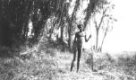 Muž s oštěpem stojící před skupinou palem v travnaté savaně.