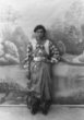 Mladá žena stojící před ateliérovým malovaným pozadím, má bohaté šperky, náhrdelníky, přívěsky u pasu, náramky