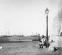 Muž sedící pod lucernou, v pozadí lodě kotvící v přístavu
