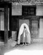 Před domem stojí žena zahalená do pláště